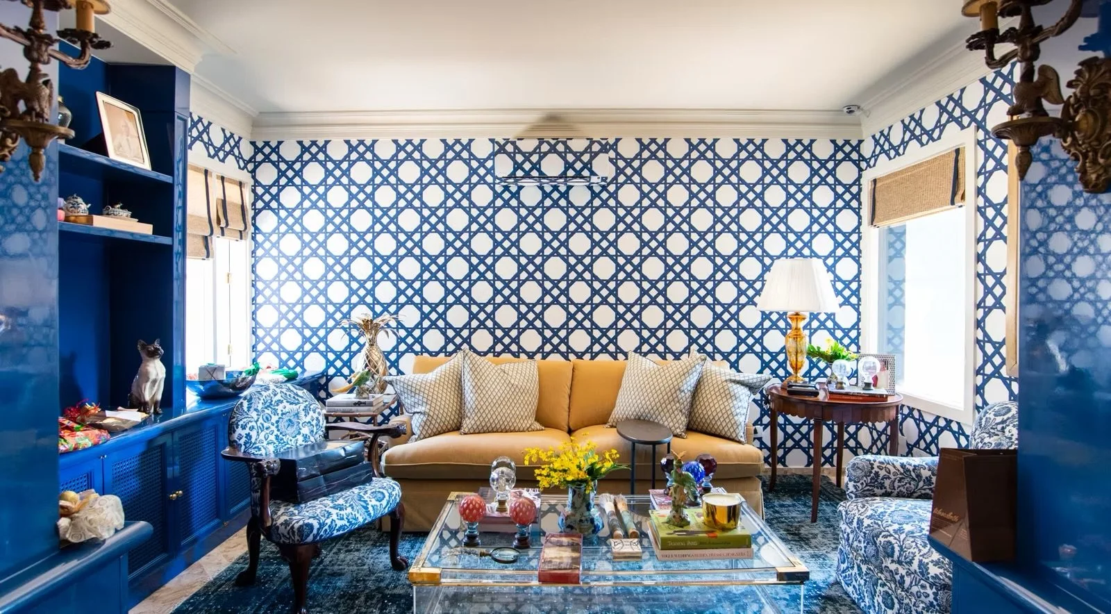 Sala de estar com parede rosa com estampa de linhas azuis formando um desenho geométrico, sofá bege com almofadas, mesa de centro de vidro com objetos decorativos e livros, poltrona de estampa azul e branca, luminária com base dourada e abajur branco, janelas com cortinas de tecido beige, teto branco com luminária no centro.