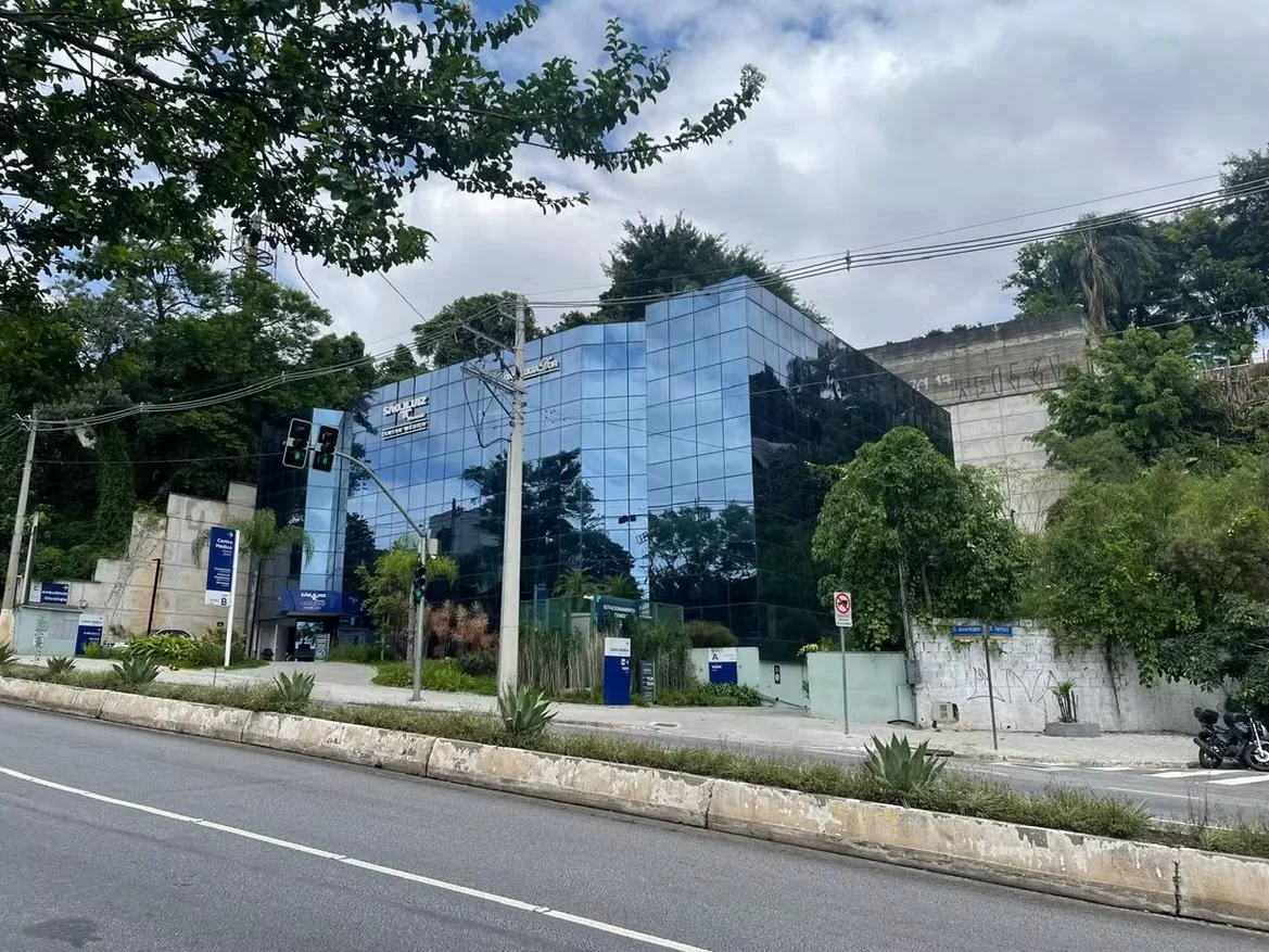 Prédio de vidro azul refletindo árvores ao redor, com sinais de trânsito, postes de energia e vegetação na frente, na cidade.