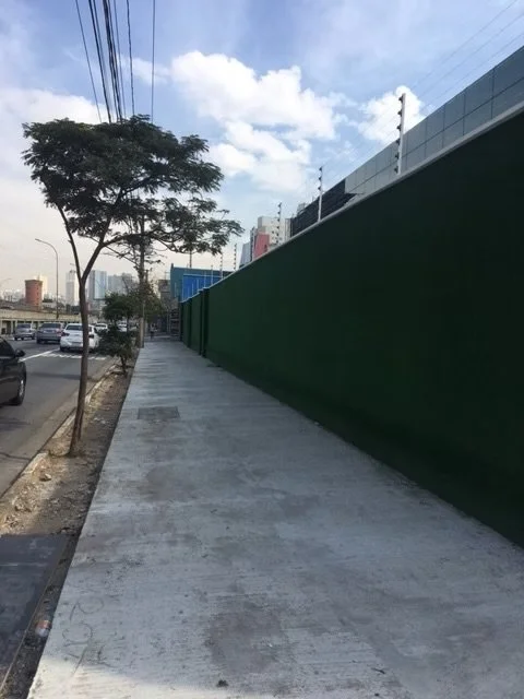 Calçada ao lado de uma rua com árvores, proteção verde e prédios ao fundo sob céu com algumas nuvens.