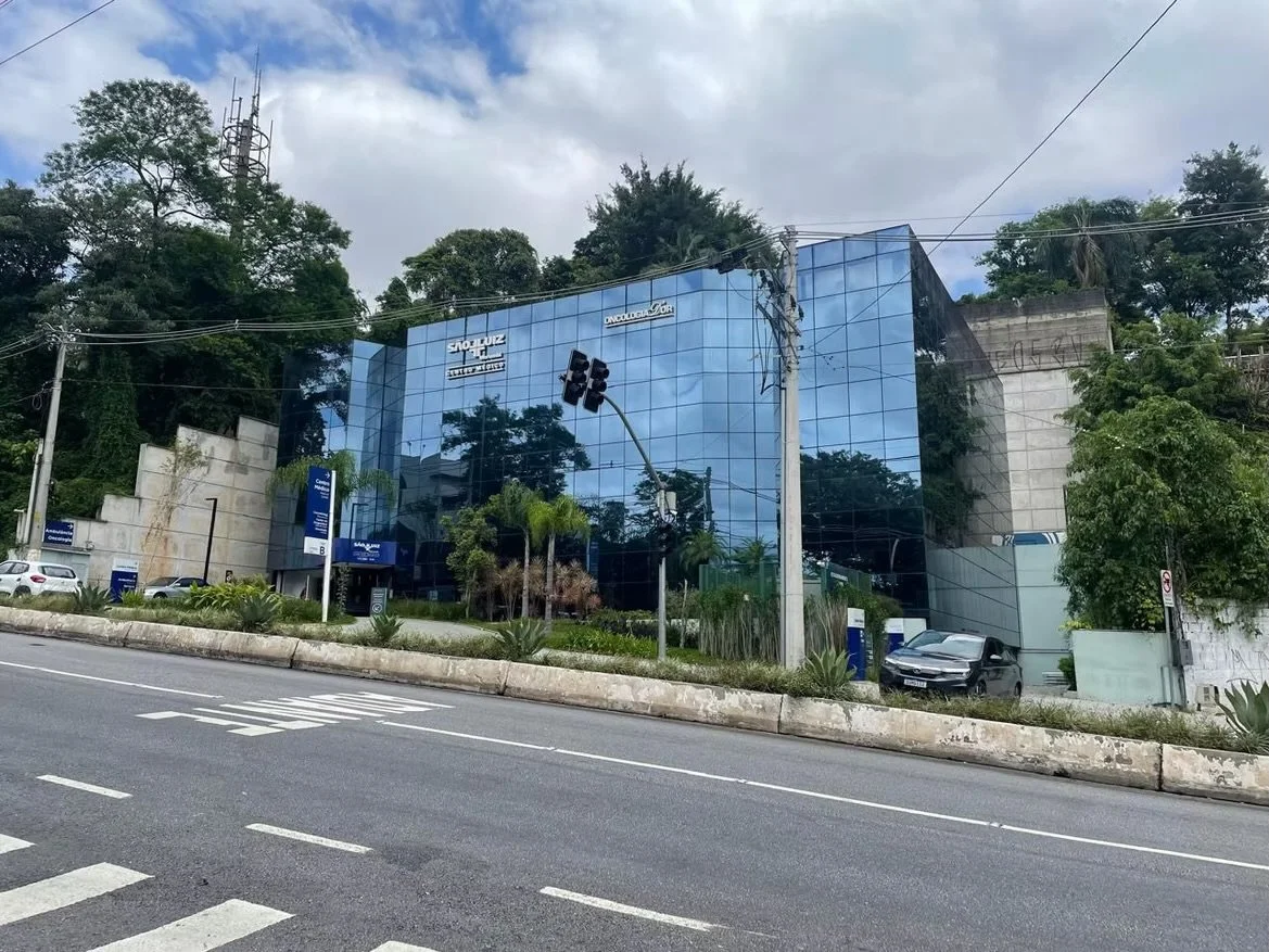 Prédio com fachada de vidro refletindo árvores ao redor, com sinais de oncologia e radioterapia e alguns veículos estacionados na frente.