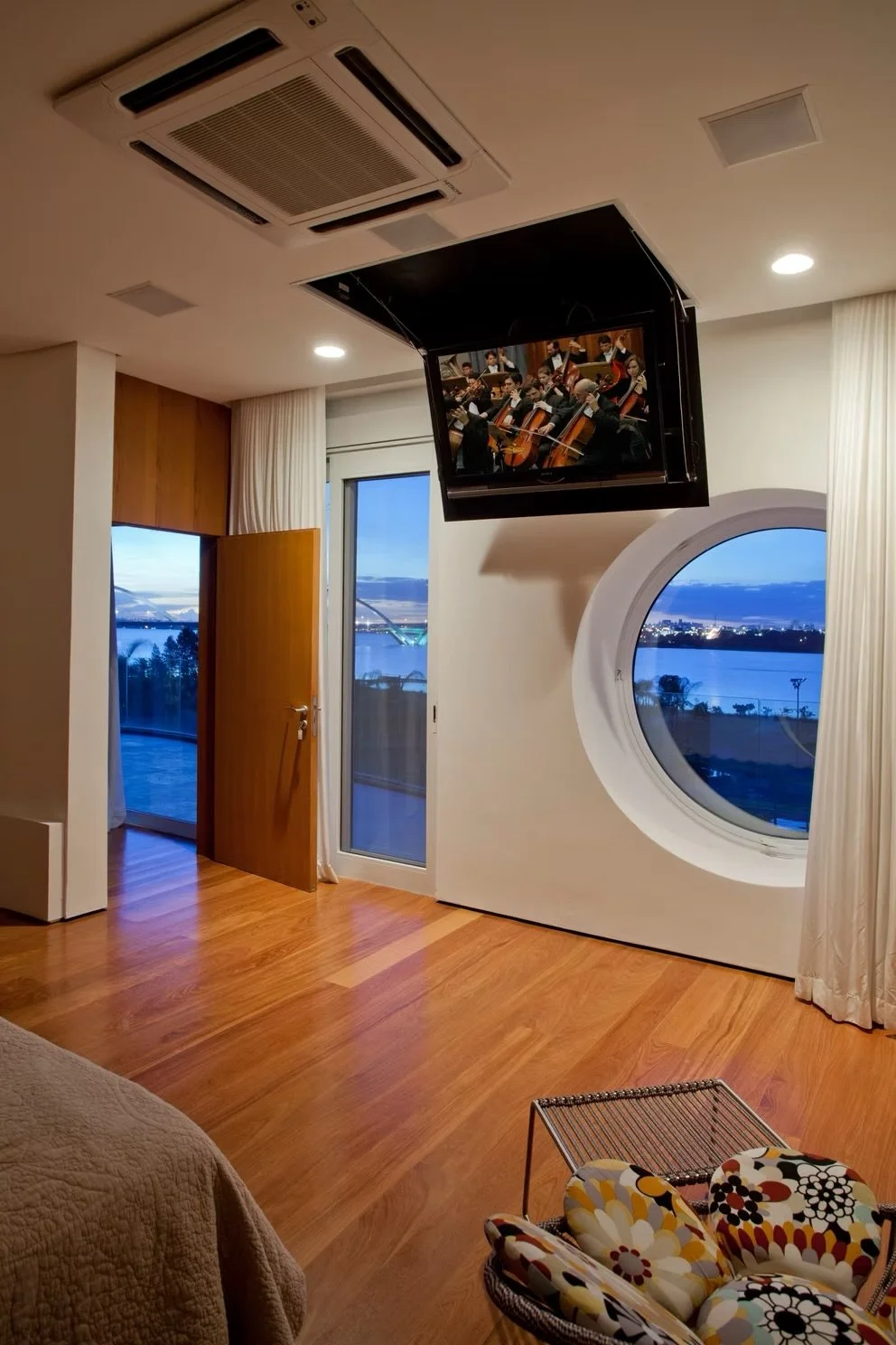 Quarto com piso de madeira, janela com vista para um lago, porta de vidro aberta, televisão ligada exibindo uma orquestra, cortinas brancas, um móvel com uma cadeira de balanço com assento florido das cores amarelo, branco, preto e vermelho, e uma pa