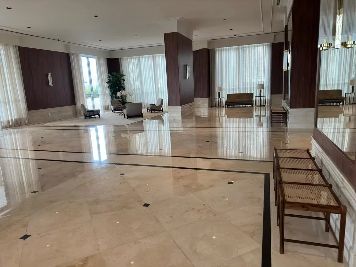 Salão de hotel com piso de mármore, paredes de madeira e cortinas brancas, com cadeiras e poltronas na decoração.