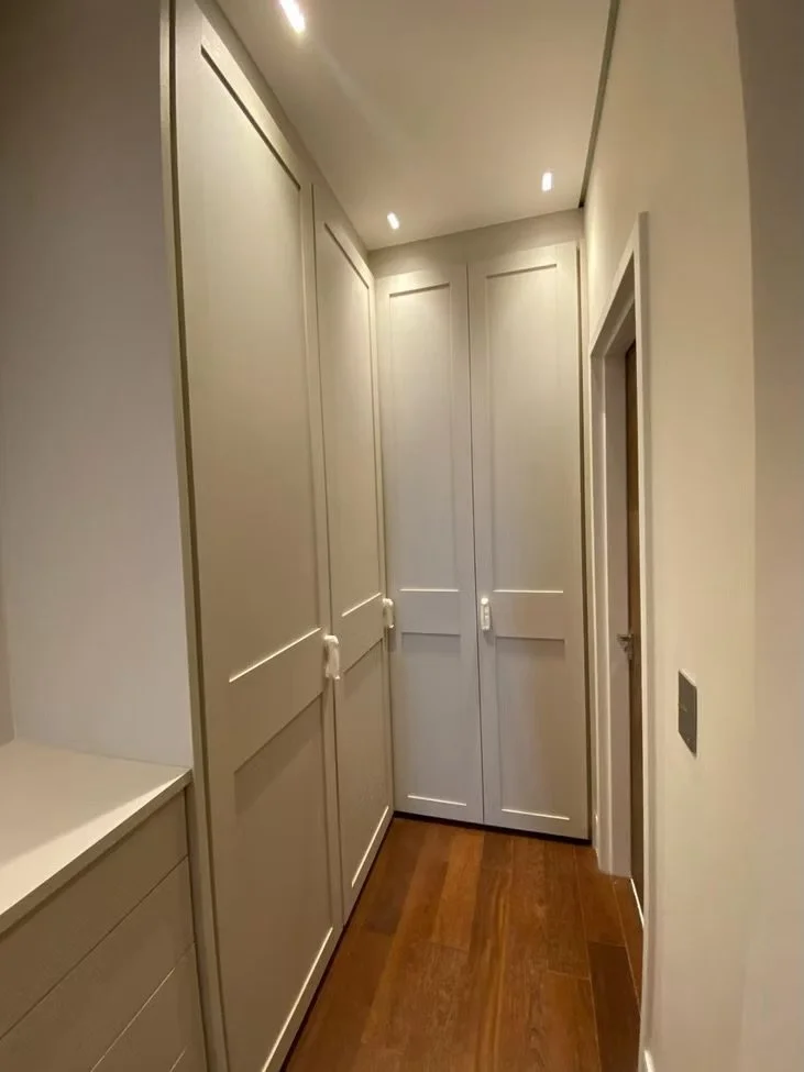 Corredor com armários embutidos brancos e portas fechadas, piso de madeira e iluminação embutida no teto.