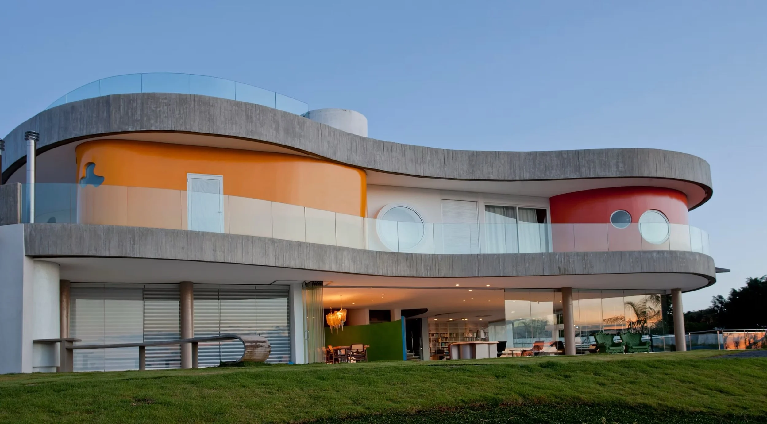 Prédio moderno com arquitetura curvilínea, com cores vibrantes de laranja, branco e cinza, com varandas de vidro e janelas circulares, cercado por um gramado verde sob céu claro.