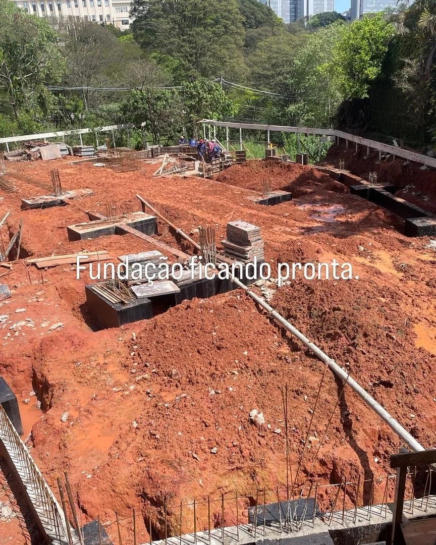 Imagem de um canteiro de obras com fundações sendo feitas, com blocos de concreto, tubulações e trabalhadores no fundo, em um ambiente ao ar livre, com árvores ao redor e prédios ao fundo. Texto na imagem: 'Fundação ficando pronta'.