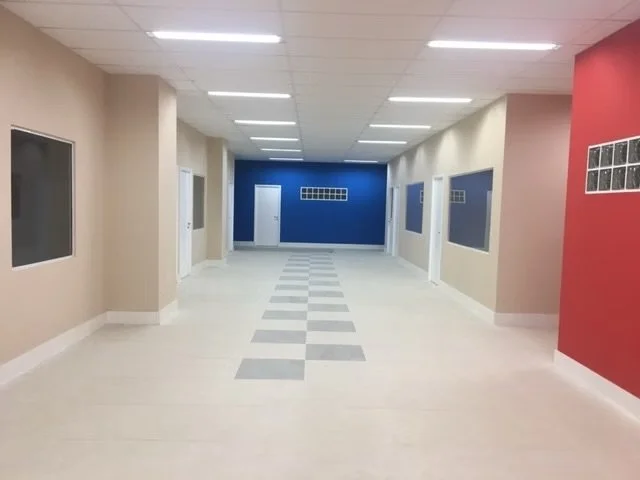 Corredor vazio com paredes em cores claras e uma parede azul ao fundo, piso quadrado e janelas nas paredes.
