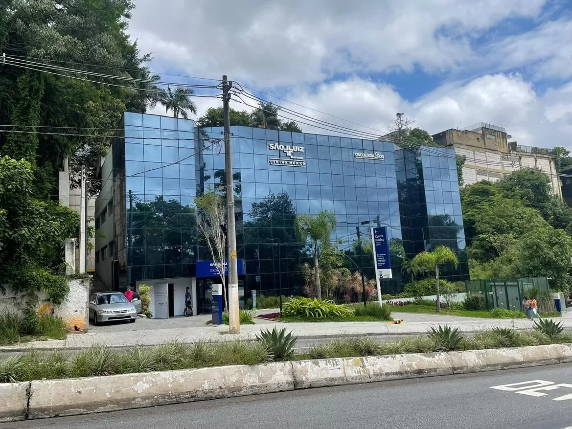 Prédio de vidro com sinais de clínicas médicas e um estacionamento na frente, árvores e céu com nuvens ao fundo.