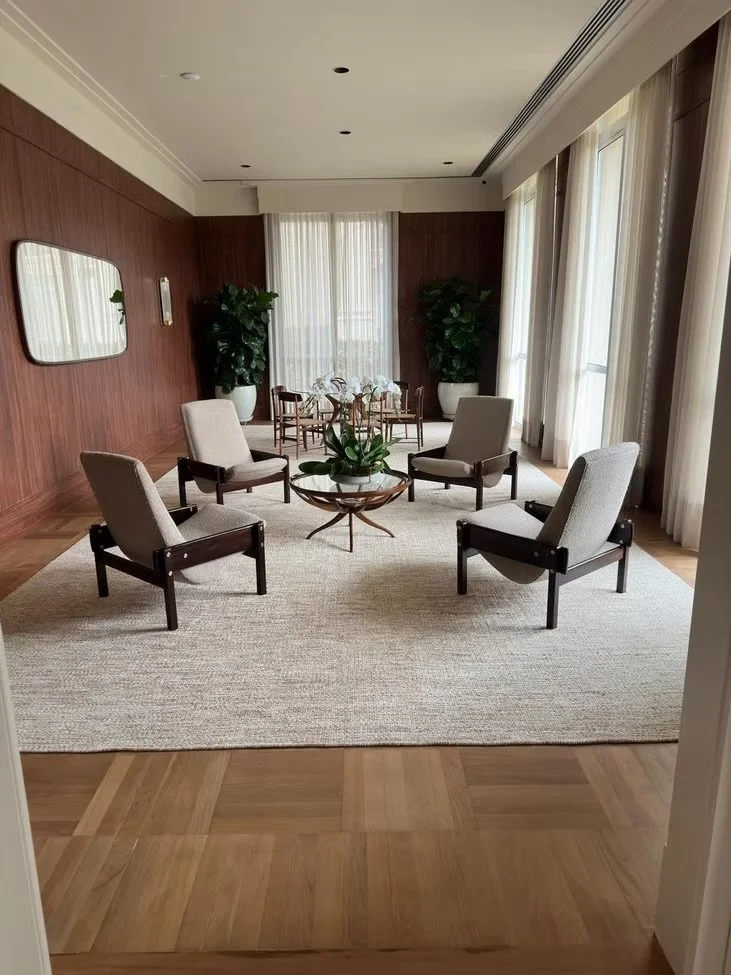 Sala de estar com paredes de madeira, janelas com cortinas transparentes, tapete claro, quatro cadeiras de encosto alto em volta de uma mesa de centro de madeira com uma planta, duas plantas grandes em vasinhos brancos ao fundo, espelho na parede à e