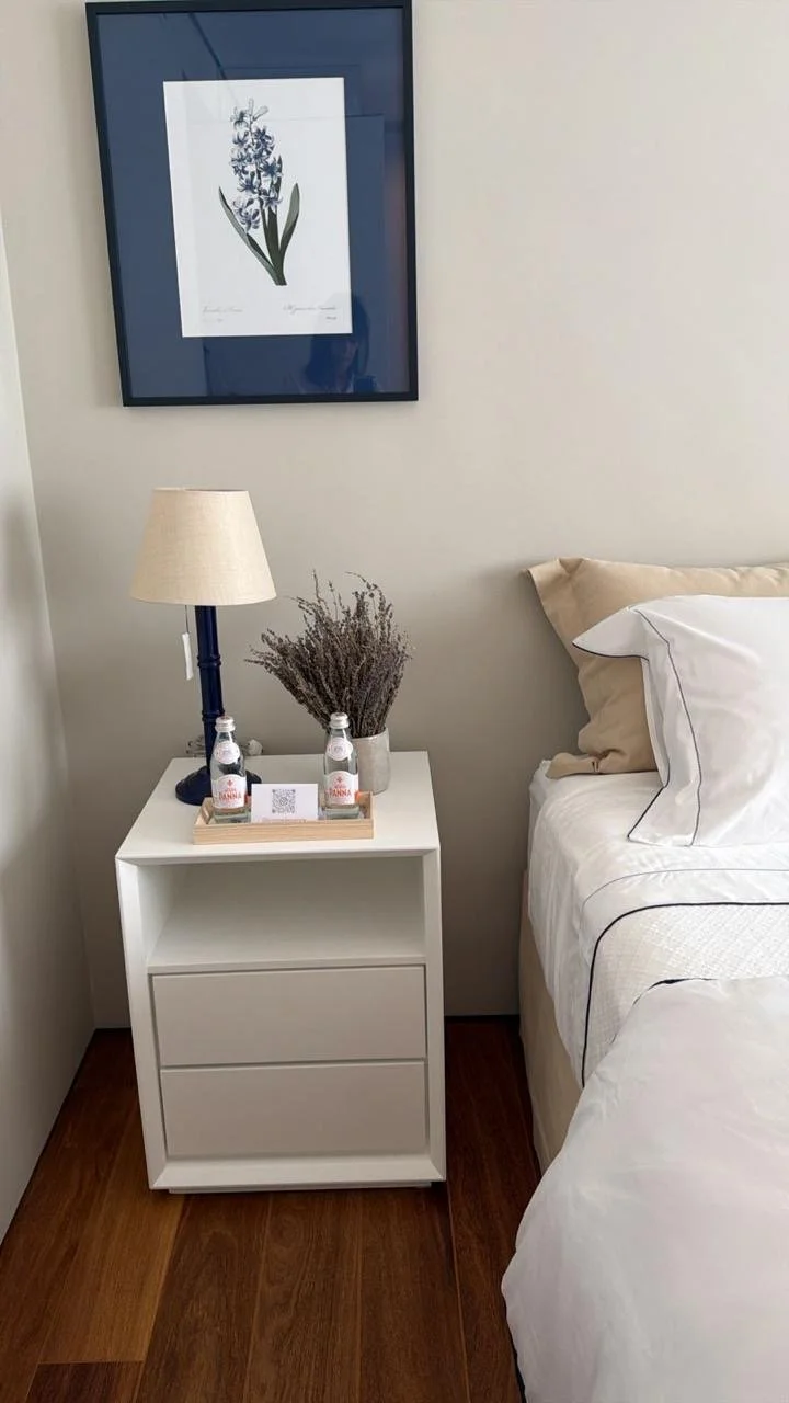Quarto com criado-mudo branco ao lado da cama, com uma luminária de abajur claro, uma planta seca, duas garrafas de água, e uma placa com QR code. Na parede, quadros de plantas, uma com moldura azul escura.