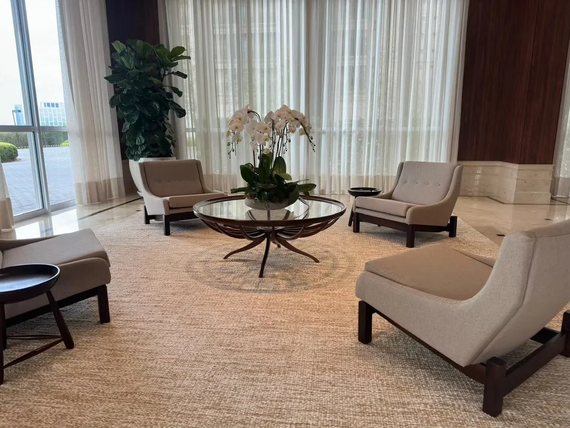 Sala de estar com poltronas brancas, mesa de centro de vidro com vaso de flores brancas, cortinas de tecido translúcido e uma planta no canto esquerdo.