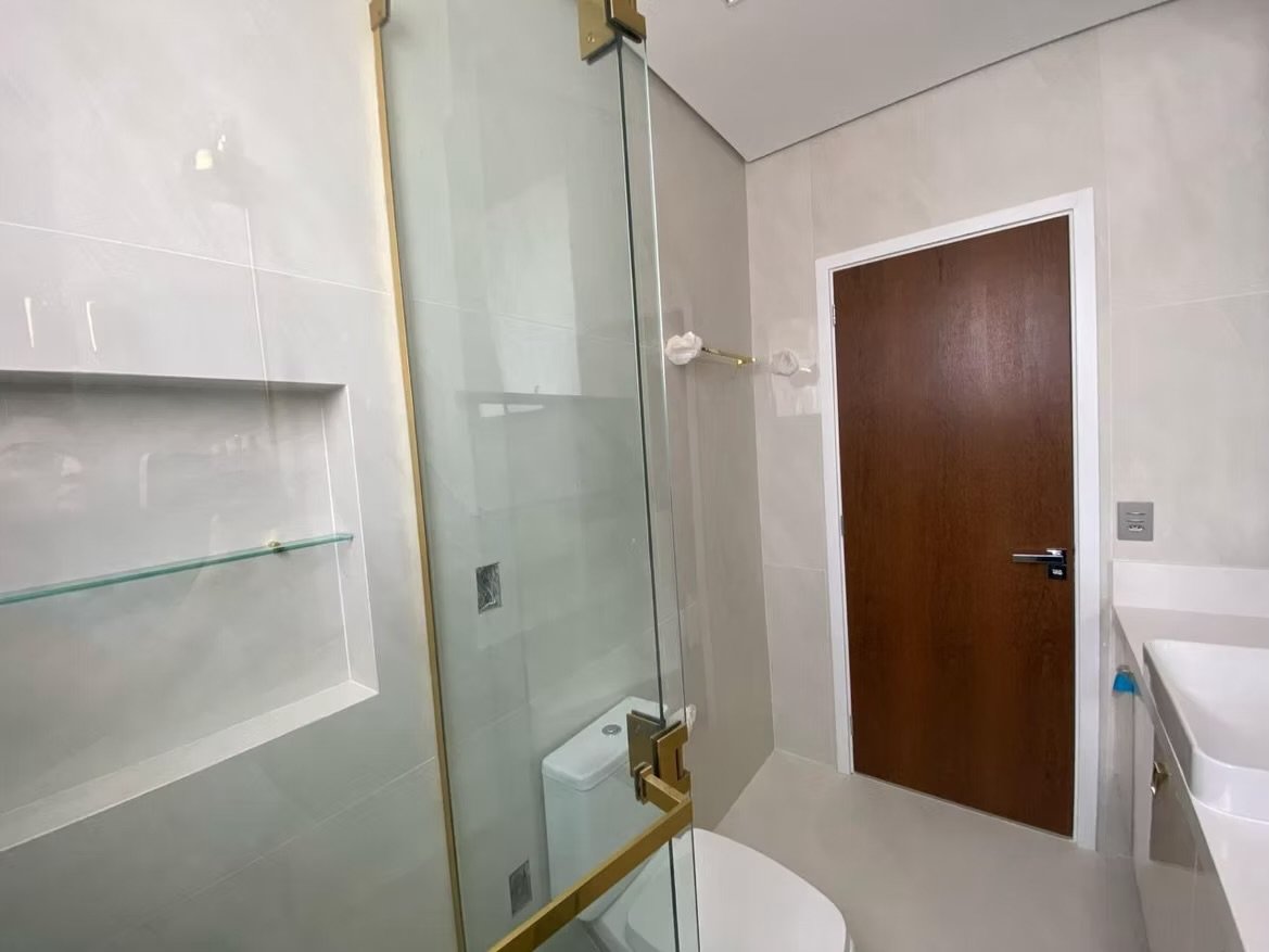 Banheiro com porta de madeira, vaso sanitário, bancada branca e box de vidro com detalhes dourados.