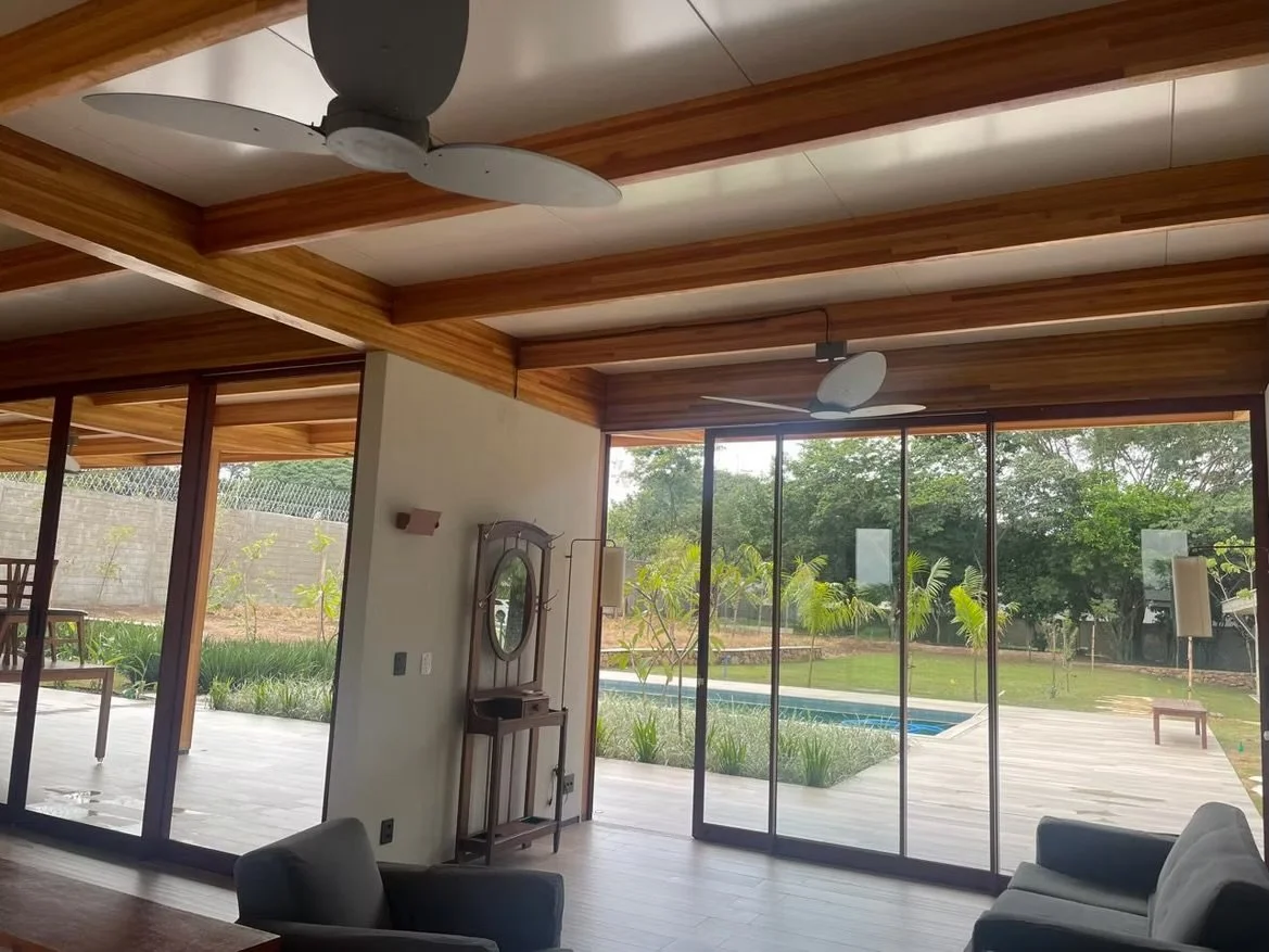 Interior de sala de uma casa com teto de madeira e ventiladores de teto brancos, paredes de vidro que mostram jardim com piscina e árvores ao ar livre, móveis de estofado cinza no interior.