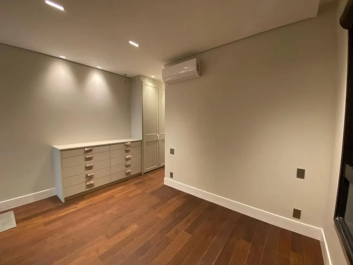Quarto vazio com piso de madeira, parede branca, ar condicionado, uma cômoda branca e uma porta.