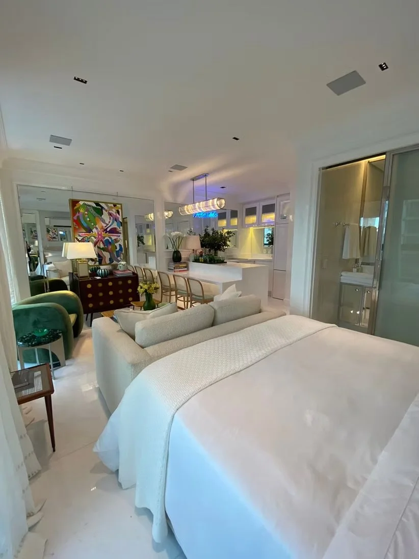 Interior de um quarto ou suíte com cama, poltronas verdes, mesa de cabeceira, decoração moderna, iluminação suave e varanda com vista para o exterior.