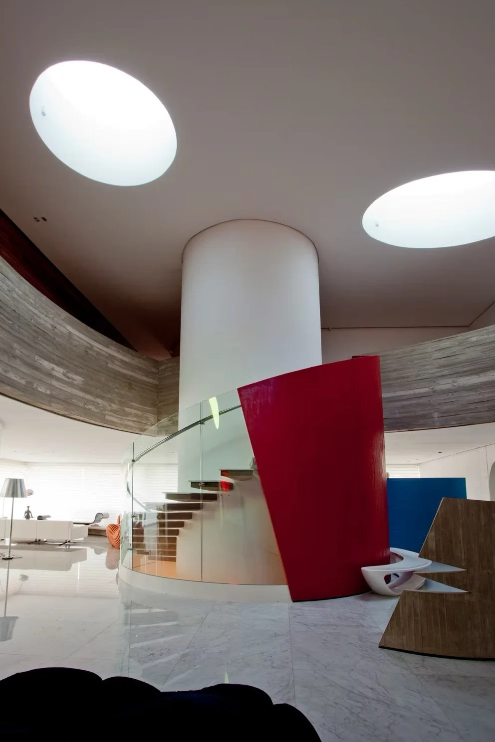 Interior moderno com escada de vidro, parede vermelha, mesa de canto branca, poltrona e luminária no fundo.