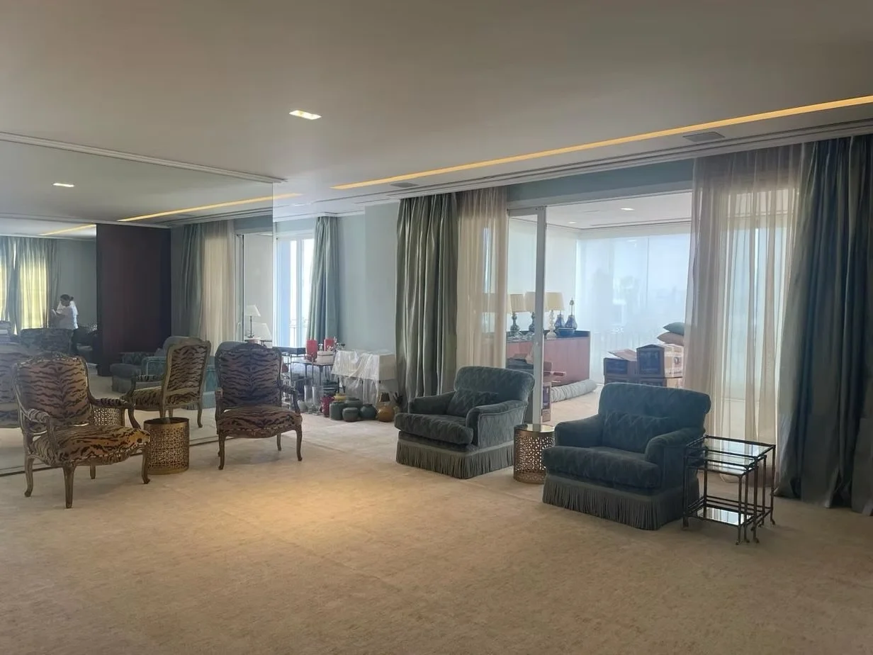 Sala de estar com diversos móveis, cortinas e uma área de varanda com janelas de vidro.