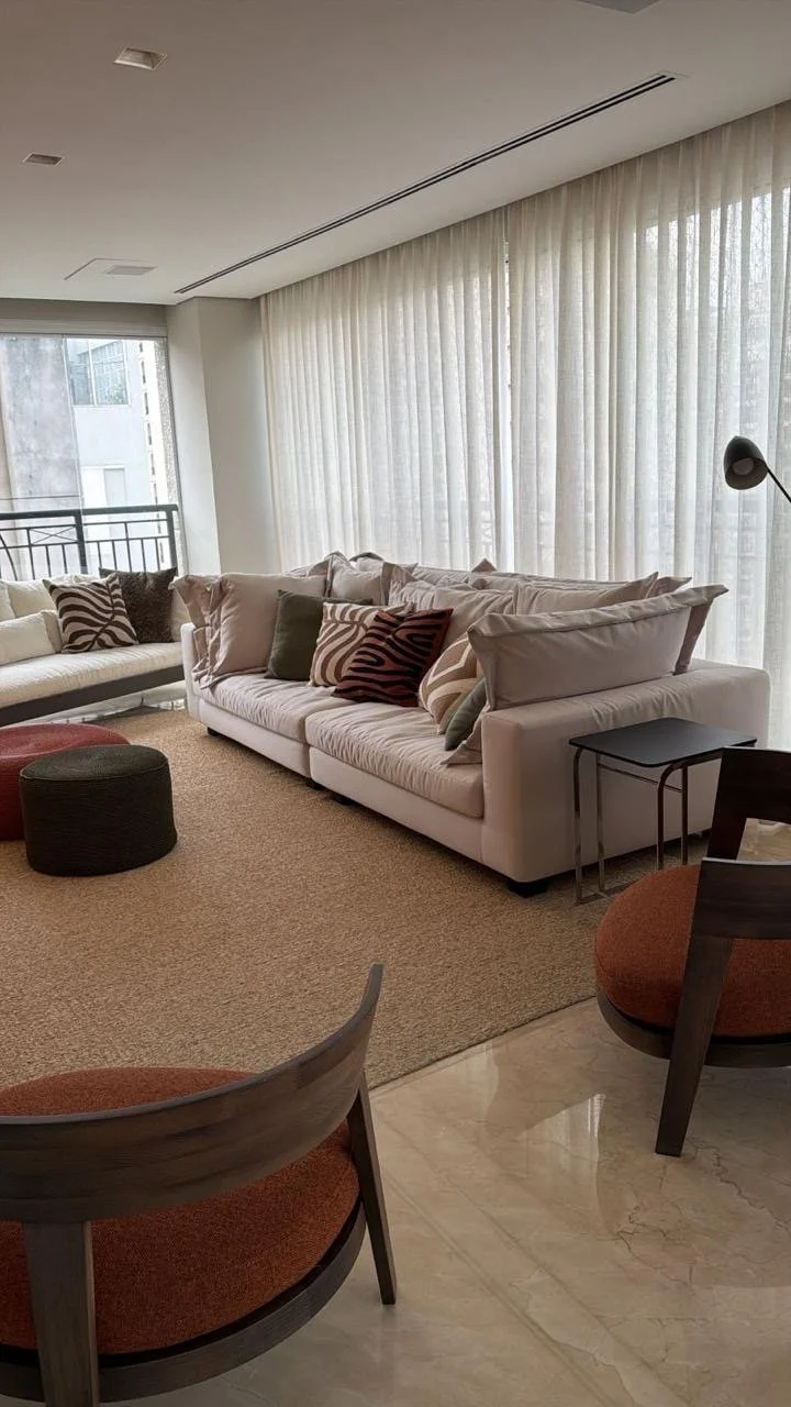 Sala de estar com cortinas brancas, sofá branco com almofadas decorativas, poltrona laranja e cadeira de madeira, mesa de centro preta, e uma área de estar com tapete bege.
