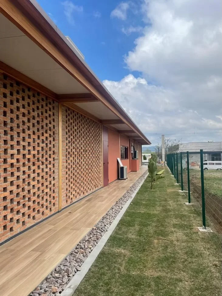 Vista lateral de uma casa com parede de tijolos de ventilação, varanda com piso de madeira, jardim com grama e cerca de metal verde.