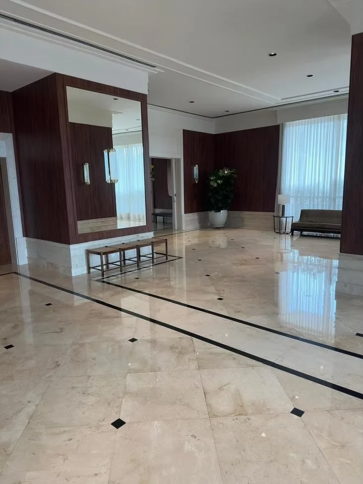 Sala de estar com piso de mármore creme, paredes de painéis de madeira escura, cortinas de tecido branco transparente, móveis simples, como uma poltrona, uma planta em vaso grande, uma luminária de mesa, e janelas grandes que deixam entrar muita luz 