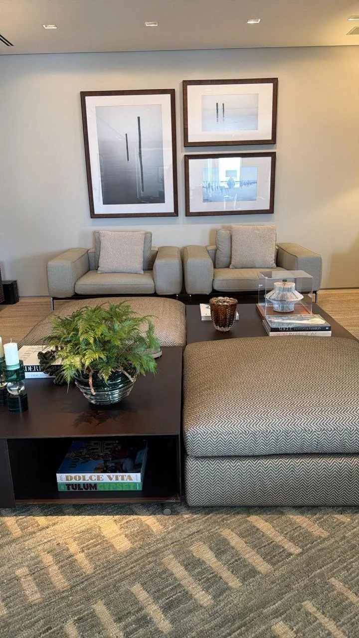 Sala de estar com sofá bege, poltrona, mesa de centro com plantas, livros e objetos decorativos, parede com três quadros abstratos.