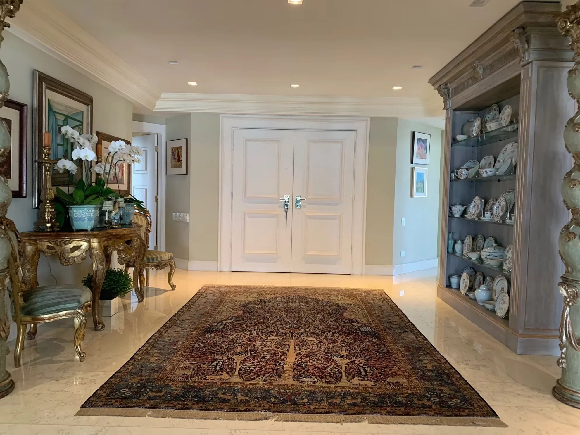 Interior de uma sala de estar com mesa de madeira, vasos de flores e quadros nas paredes. Ao fundo, porta e uma estante com louças decorativas, tapete e iluminação embutida no teto.