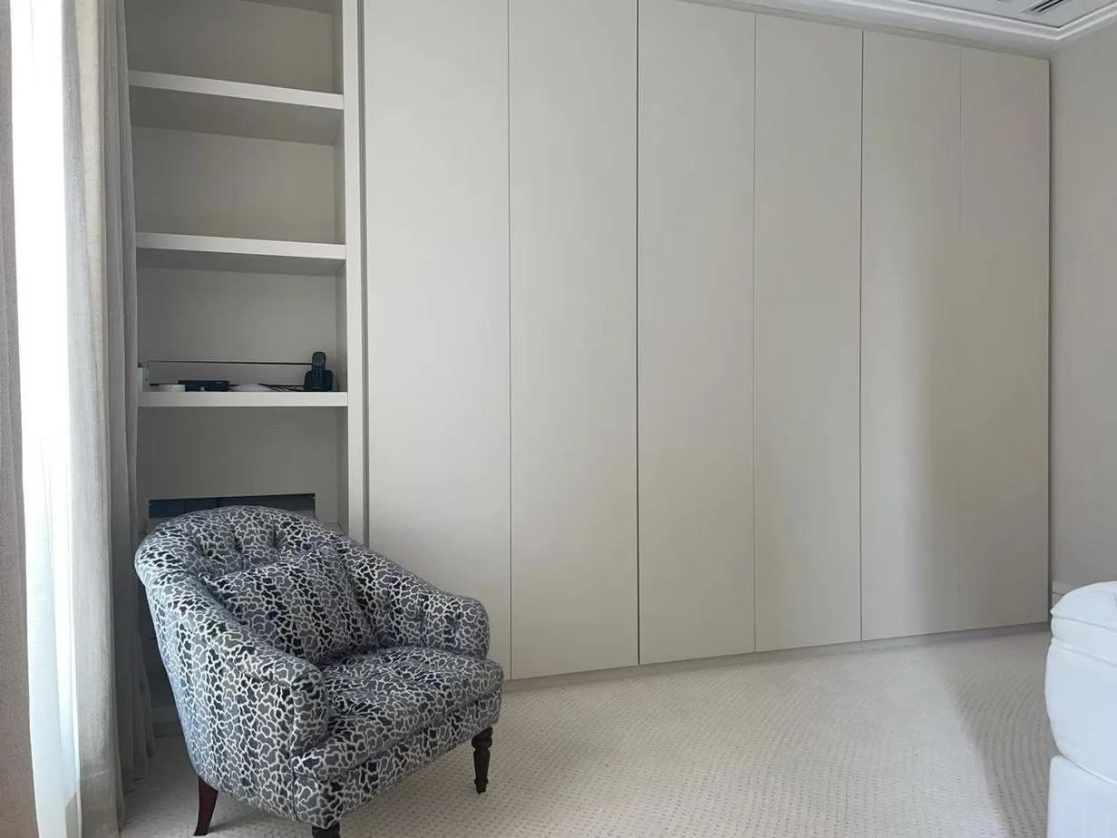 Sala com armário de portas brancas, uma cadeira de braço com estampa de leopardo, cortinas cinzas e piso de carpete claro.