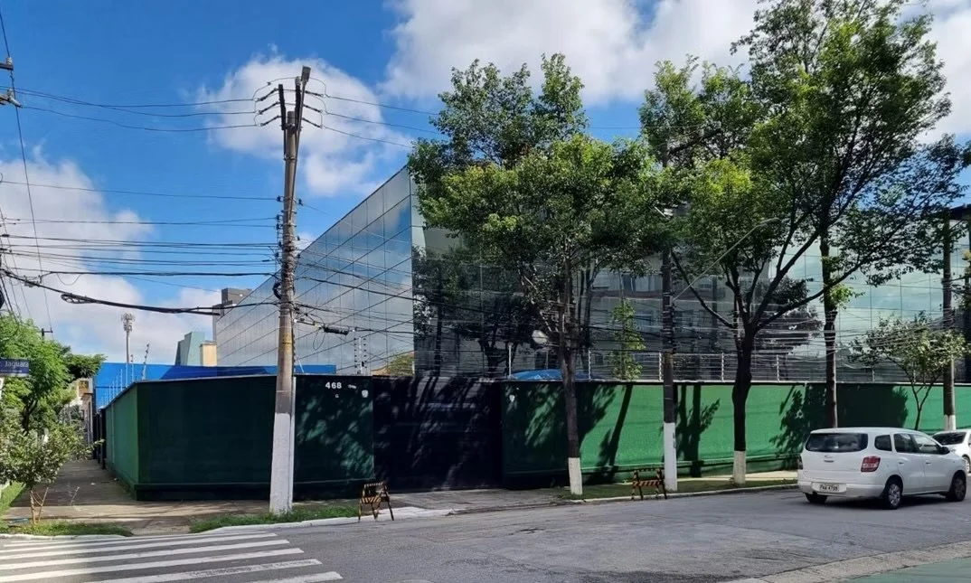 Rua com árvore, postes de energia, carro branco, edifício de vidro e muro verde