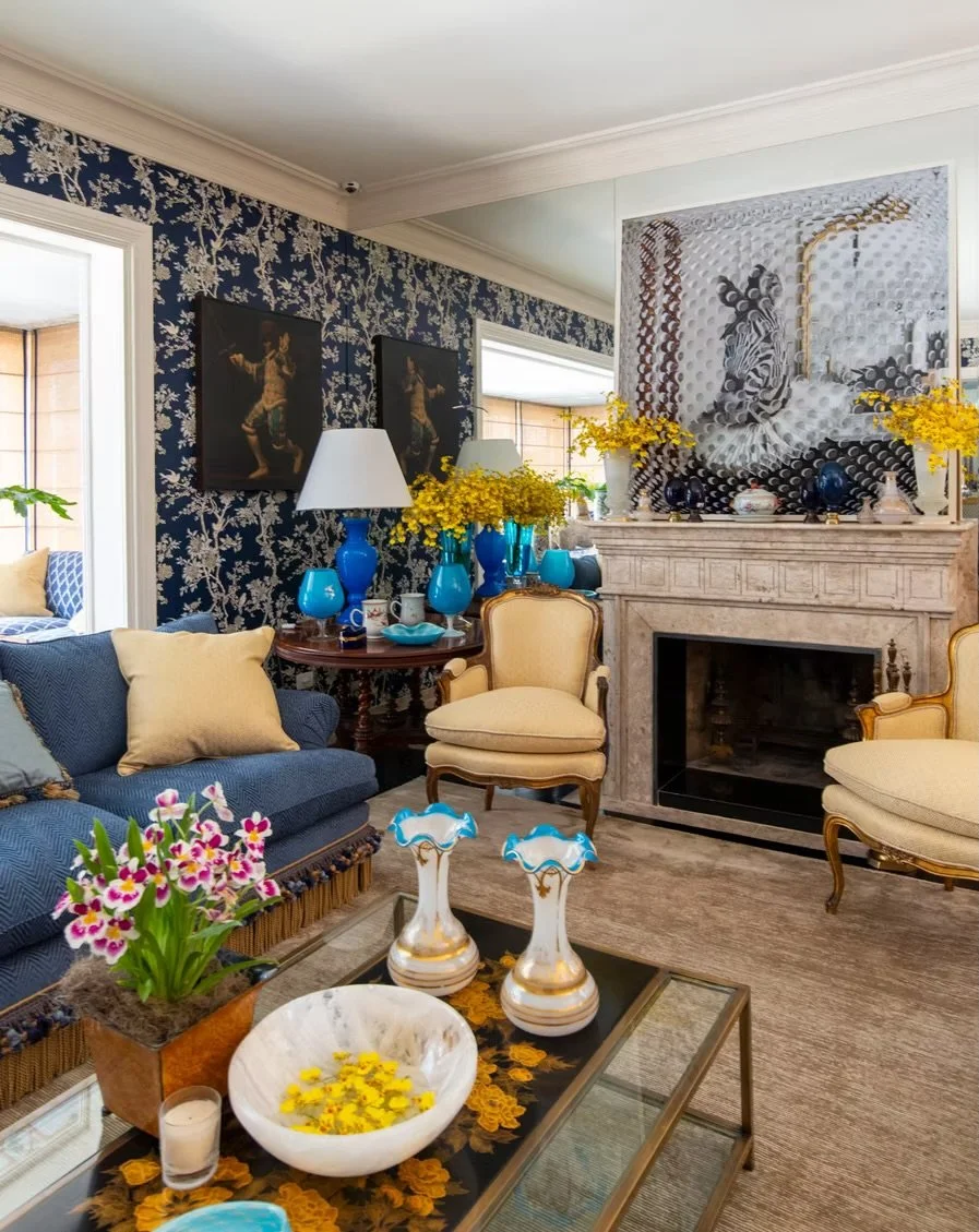 Sala de estar decorada com poltrona amarela, sofá azul, mesa de centro com vasos decorativos e buquê de flores. Paredes com papel de parede floral azul e branca, e quadros na parede. Chaminé de mármore com vasos e flores amarelas. Pintura abstrata mo