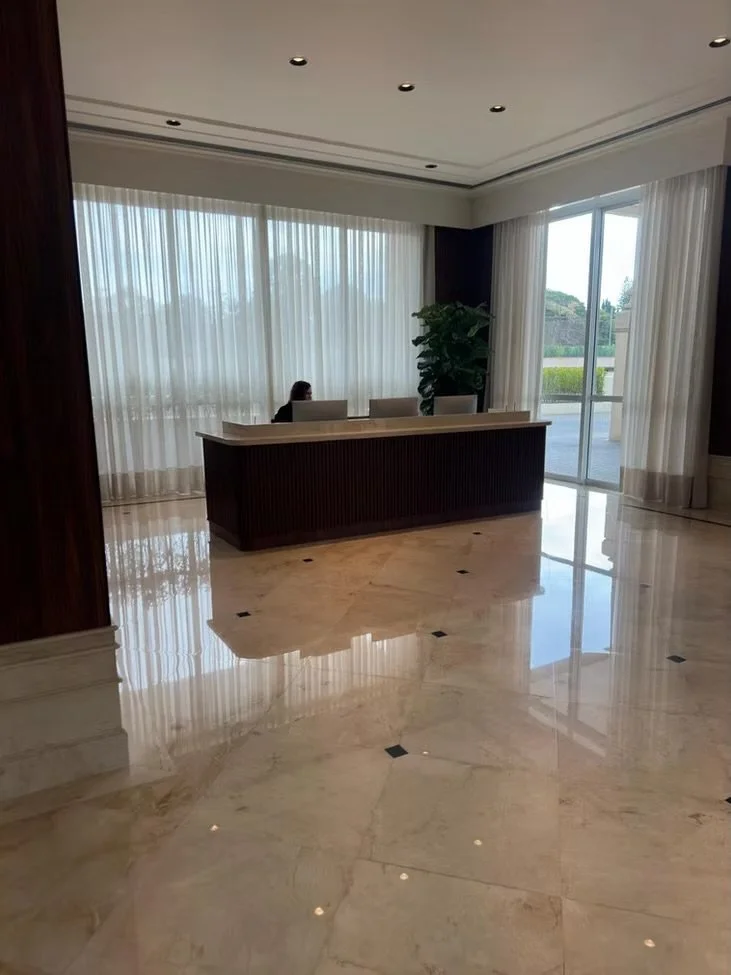 Recepção de um prédio com uma escrivaninha, uma pessoa sentada atrás dela, cortinas brancas, uma planta grande ao lado da janela e piso de mármore.