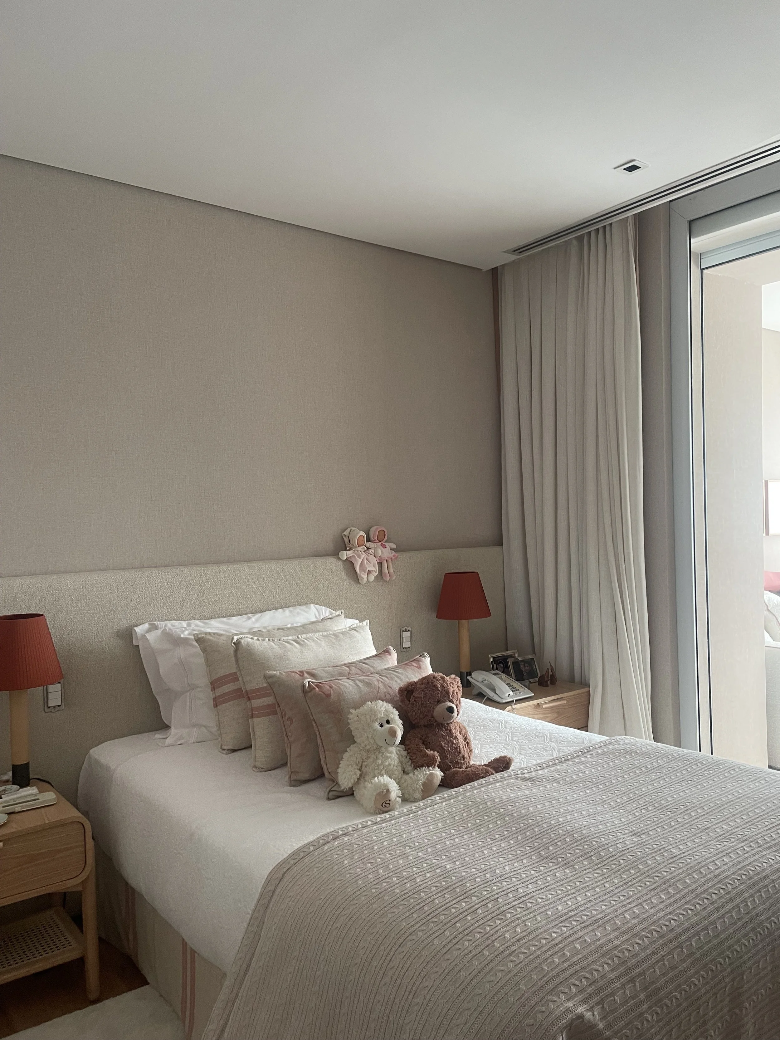 Cama de casal em um quarto com cabeceira almofadada, com dois ursinhos de pelúcia sentados na cama. Ao lado da cama há uma mesa de cabeceira com uma luminária de abajur vermelho, um telefone e alguns objetos. A parede atrás da cama é branca e há cort