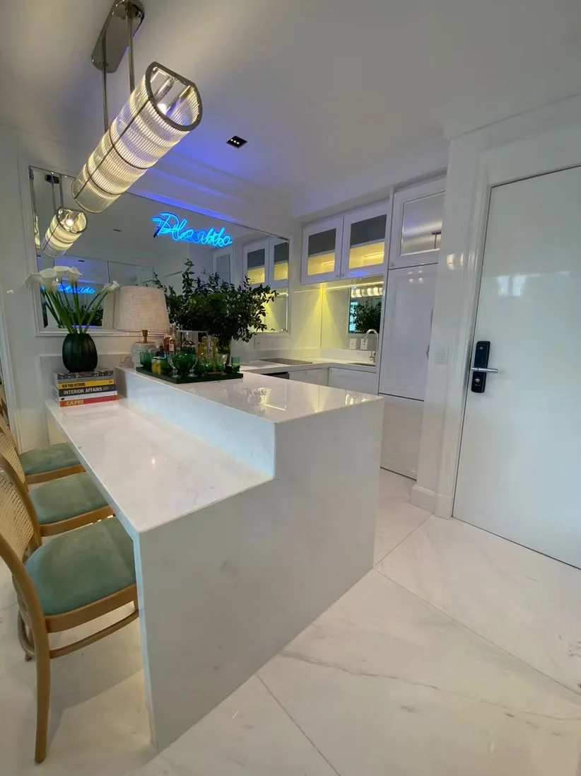 Cozinha moderna com balcão de mármore branco, decorada com plantas, livros e luminária. Espelhos na parede refletem o ambiente com uma neon azul que diz "Palesso".