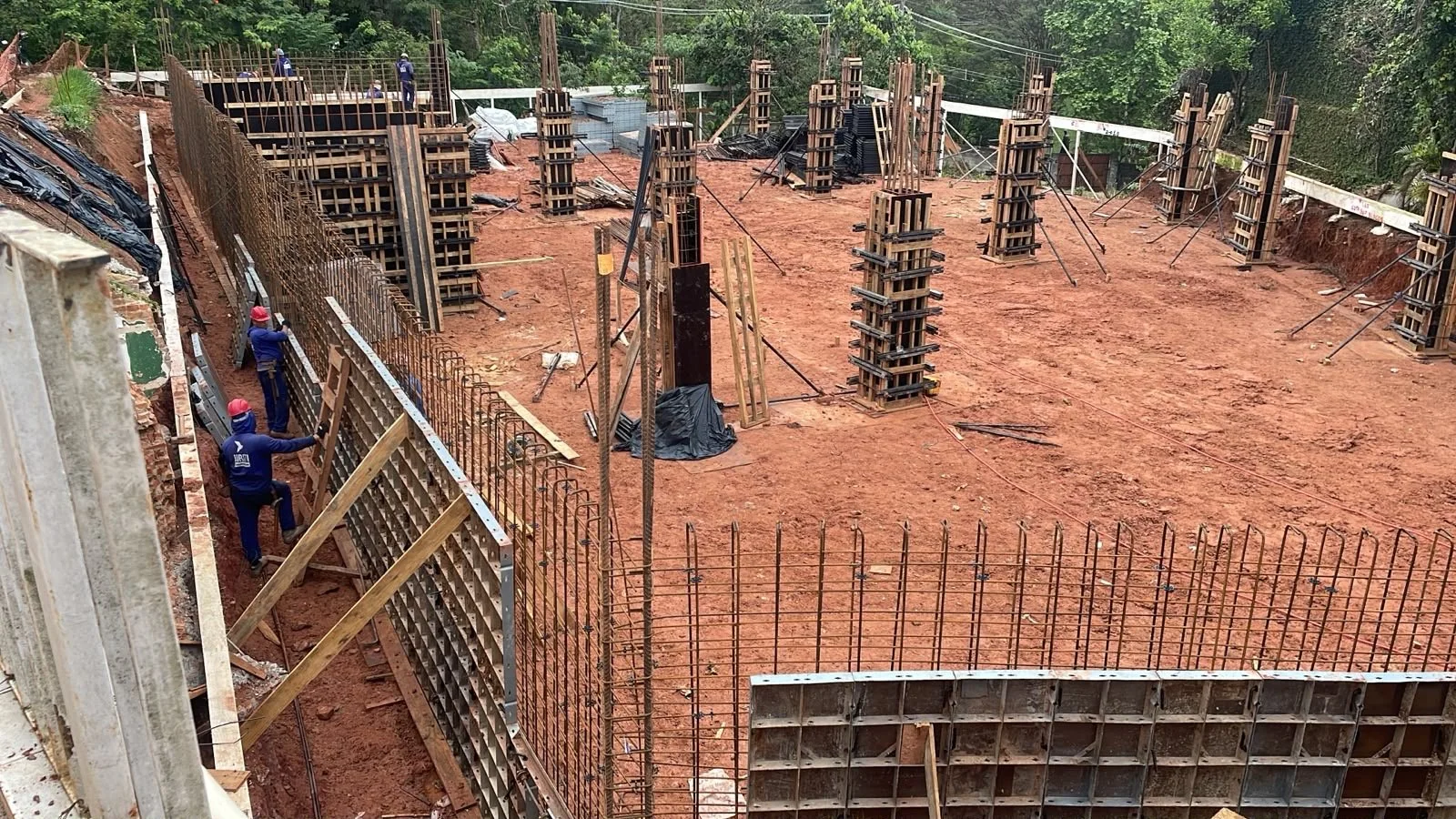 Construção de fundações com formas de madeira e ferros em um terreno de terra batida, com trabalhadores usando capacetes vermelhos.