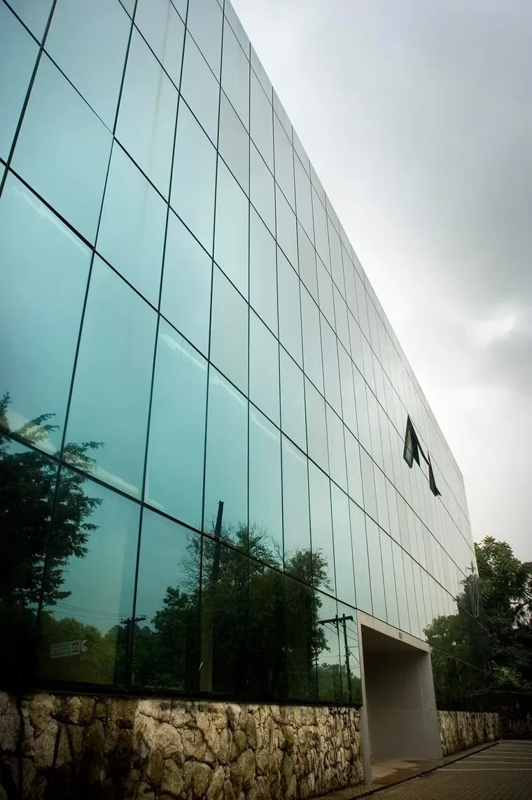 Fachada de um edifício moderno de vidro refletindo o céu e árvores ao redor, com estrutura de pedra na base