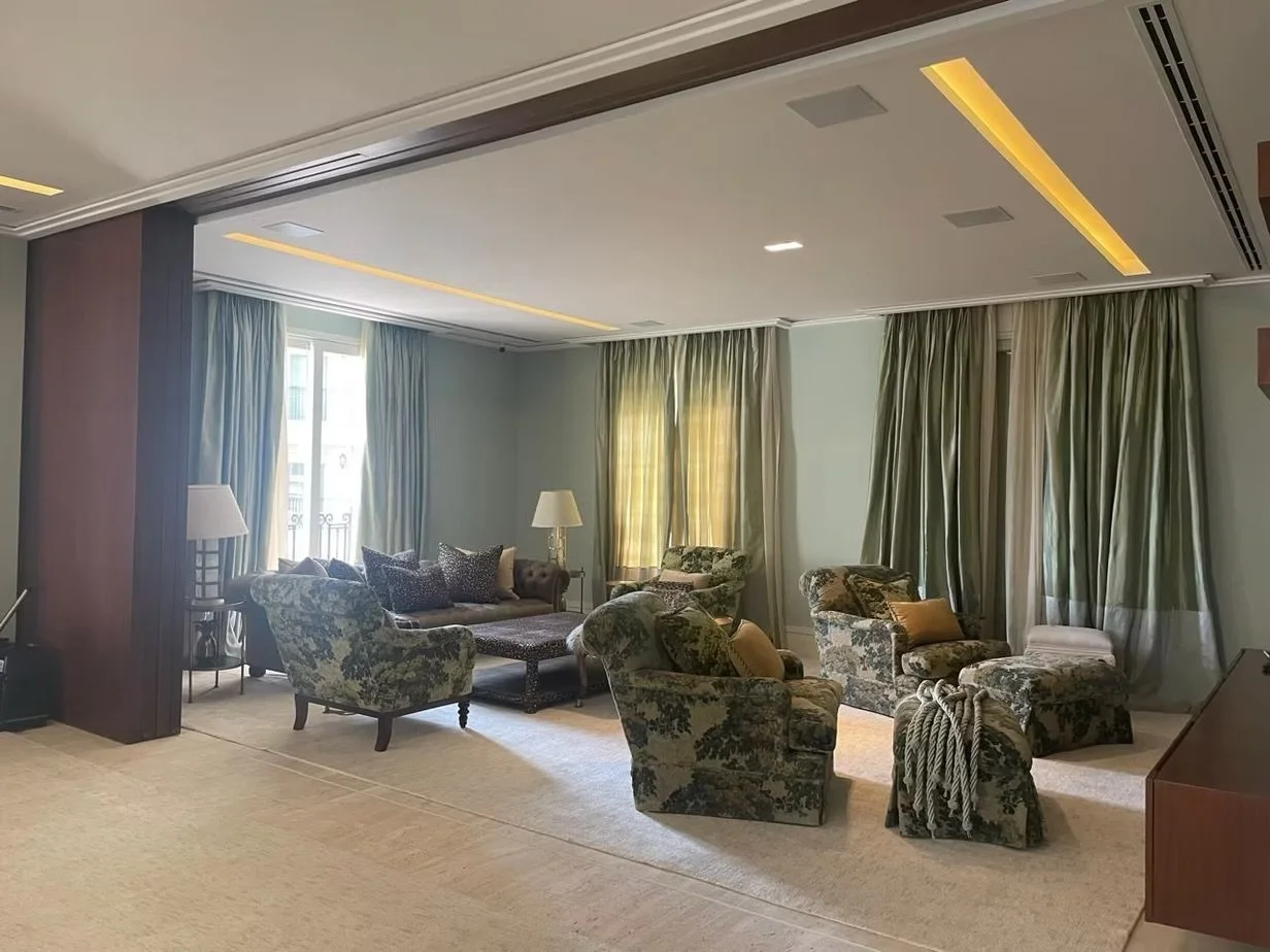 Sala de estar com poltronas e sofá com estofado floral, cortinas verdes e amarelas, luminárias de mesa, piso de carpete claro e teto com iluminação embutida.