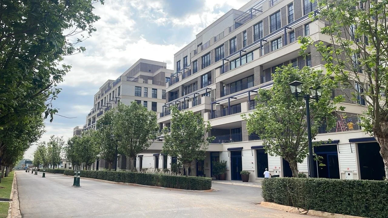 Rua com árvores e edifícios residenciais modernos com varandas, ao dia com céu parcialmente nublado.