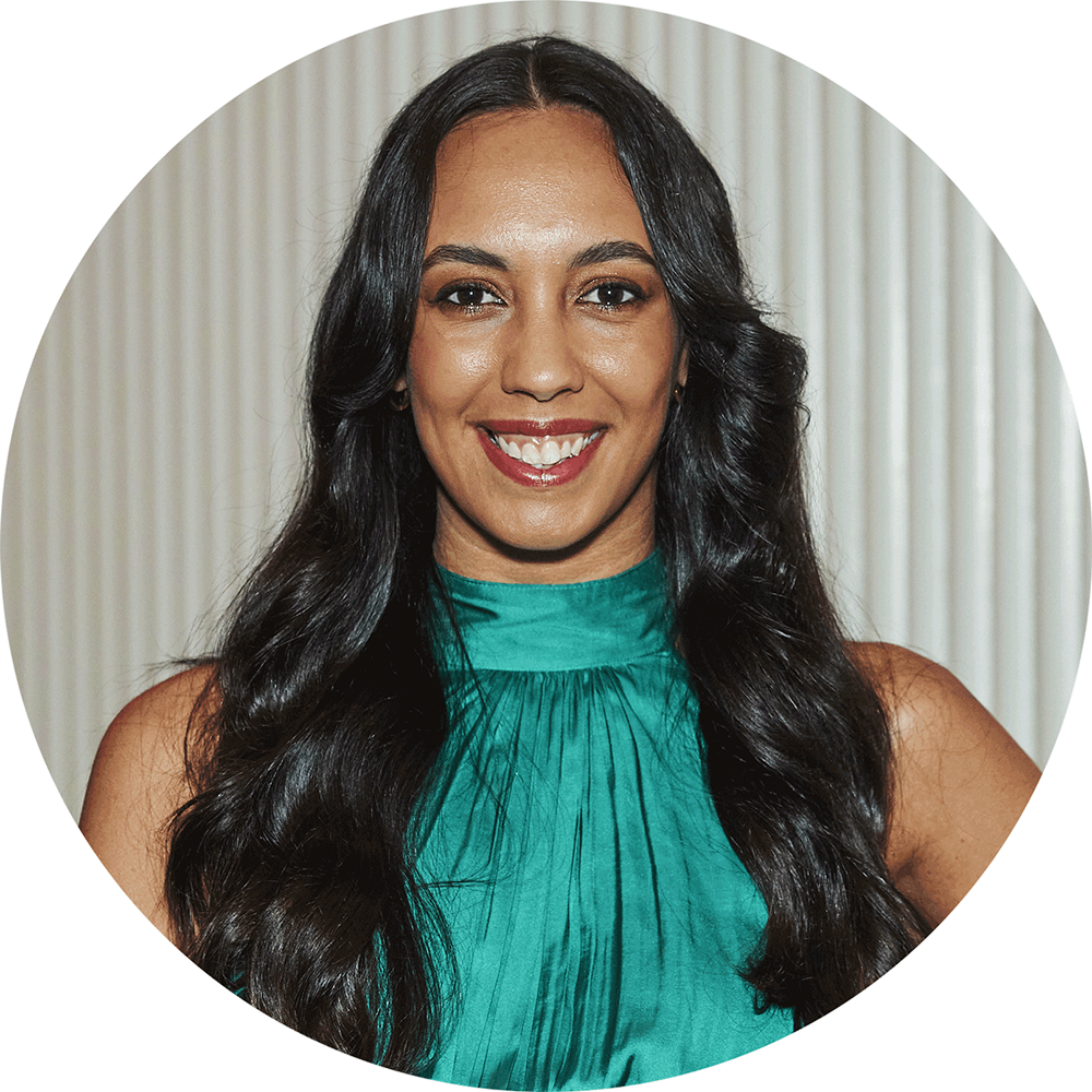 A woman with long black hair smiling, wearing a teal sleeveless top, standing in front of a white curtain.