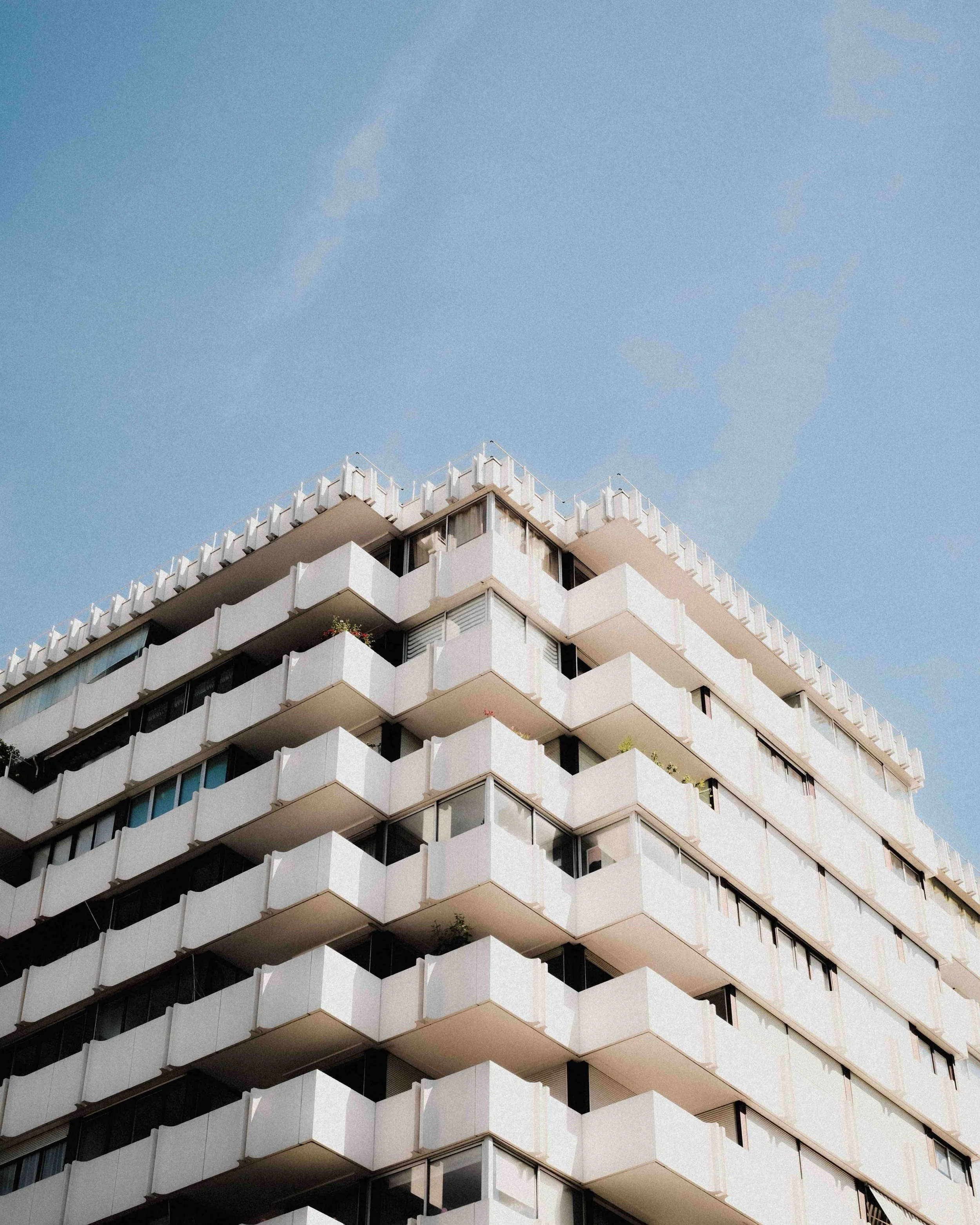 A multi-story white modern apartment building with balconies and floor-to-ceiling windows, under a clear blue sky.