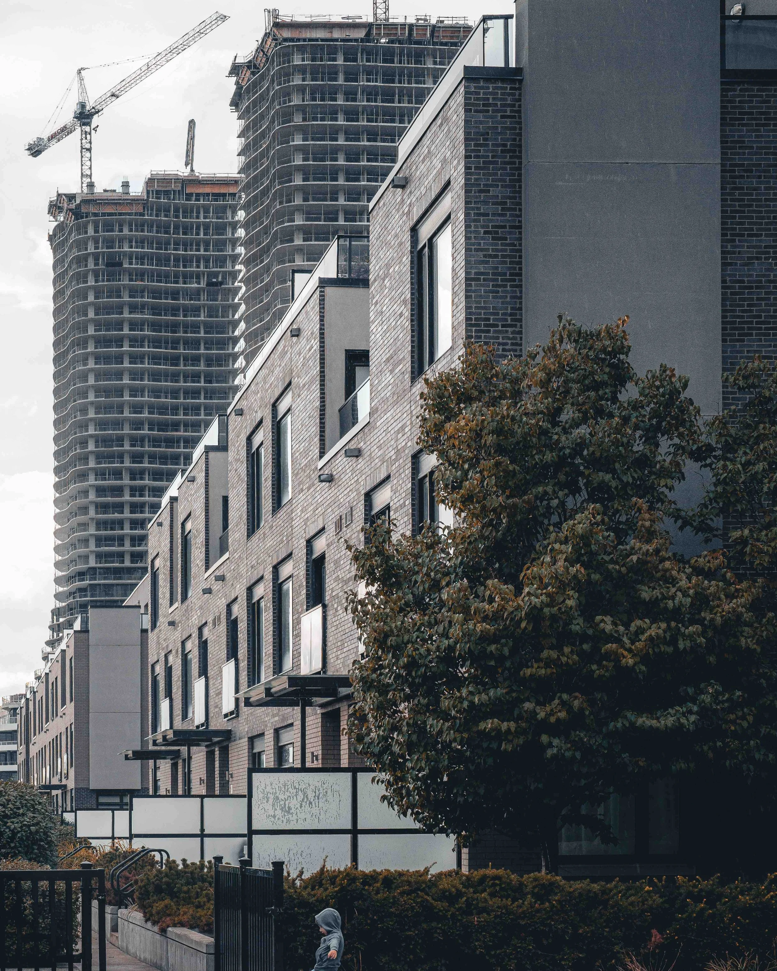 A cityscape with high-rise buildings, a construction site with cranes, and a small child in a hoodie walking along a sidewalk in a residential area with trees and greenery.