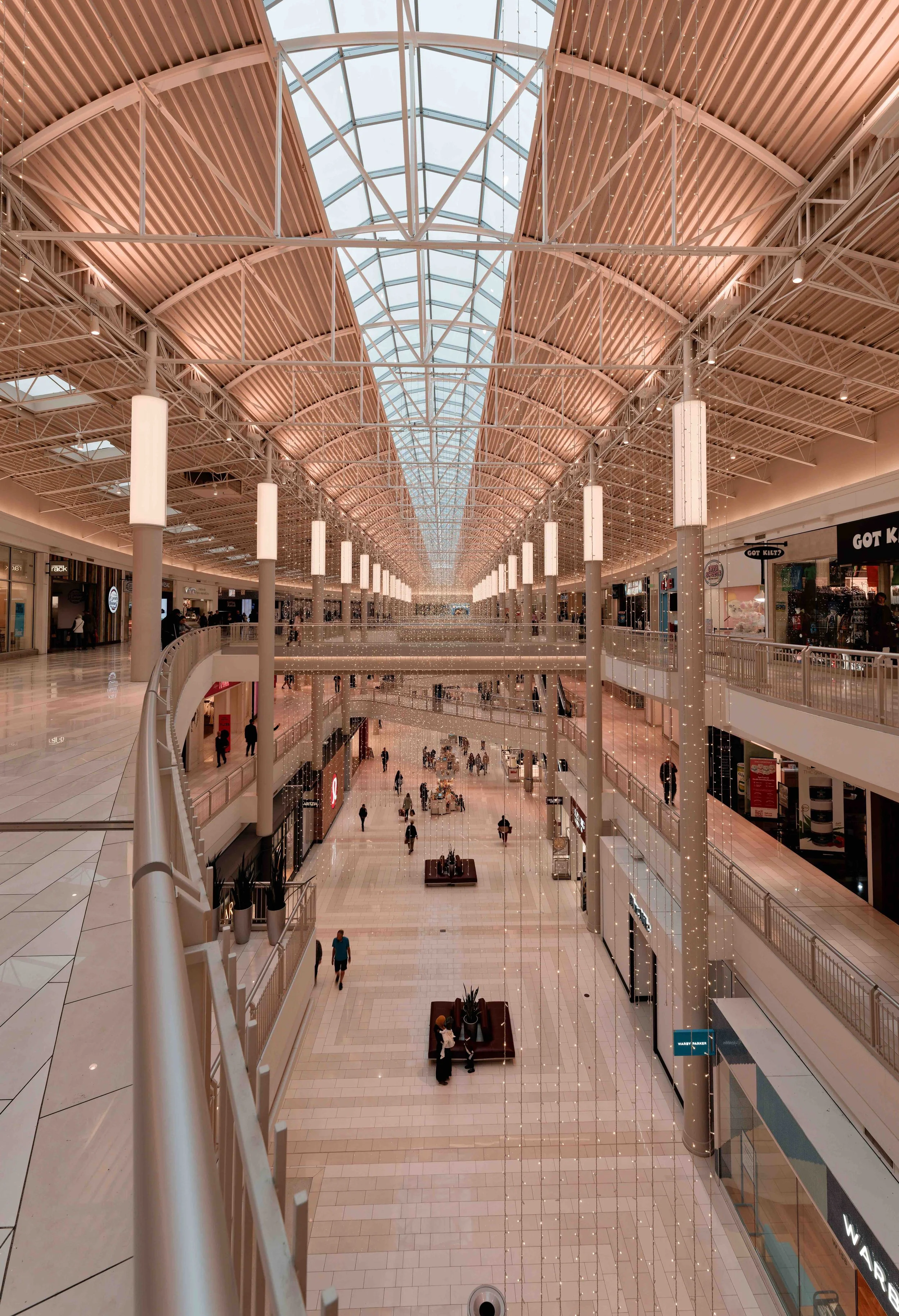 Interior view of a modern, multi-level shopping mall with a curved glass roof and various stores along the upper and lower levels. Pedestrians walk on tiled floors with railings and lighting accents.