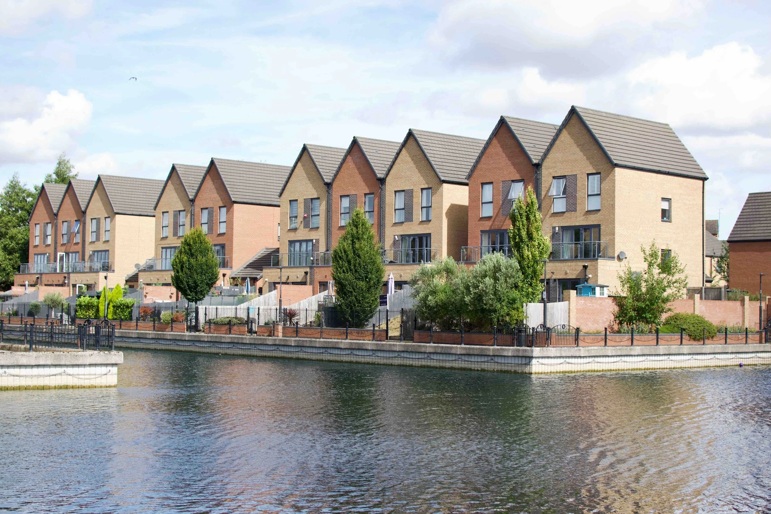 Row of modern townhouses with brick facades and balconies, situated along a body of water with trees and a cloudy sky in the background.