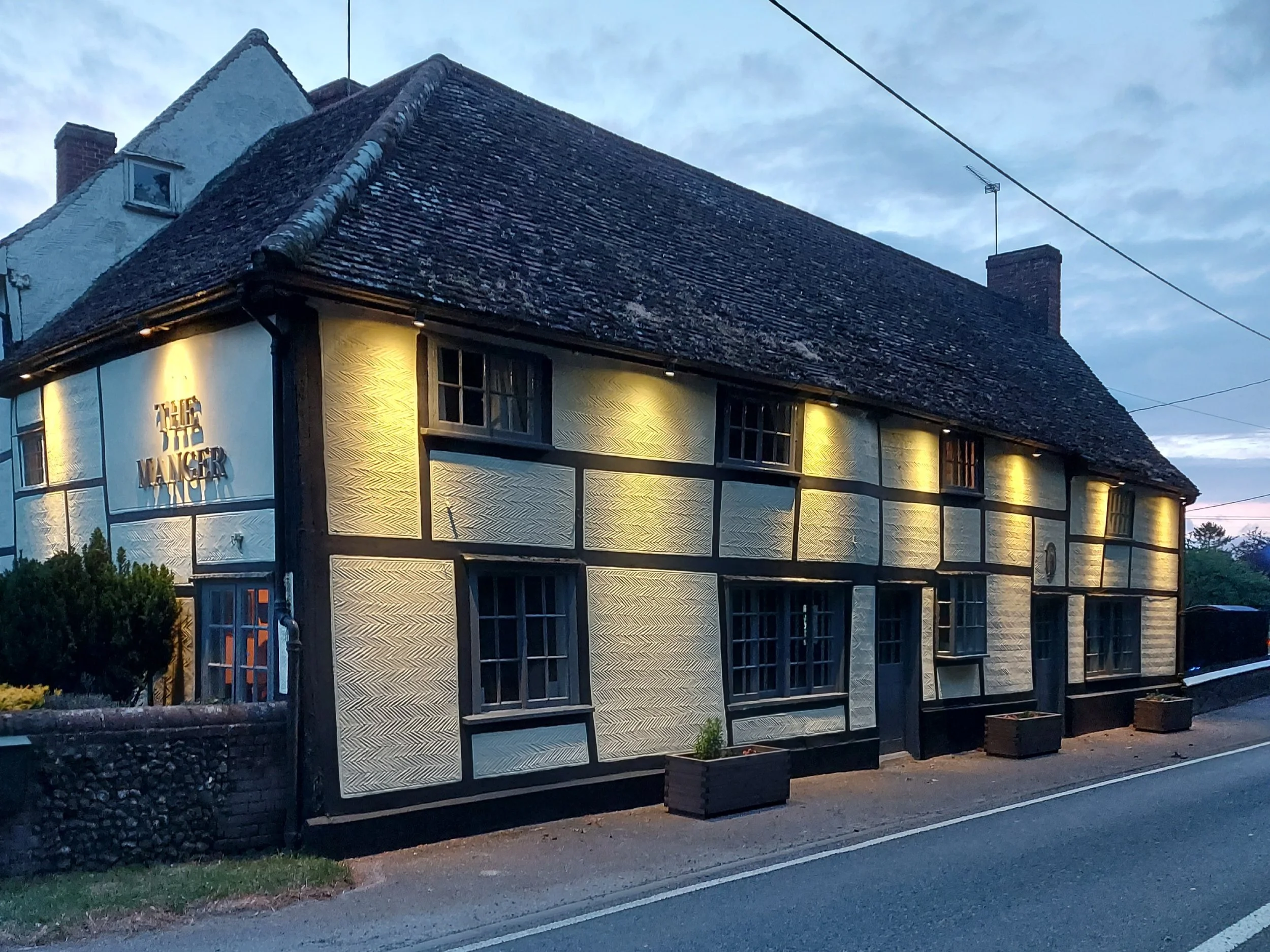 A building with a sloped roof, illuminated by exterior lights, with a sign that reads 'The Warner'. The building has a black and white half-timbered design, with several windows and a chimney. There are small planters at the sidewalk edge, and power lines overhead, with a partly cloudy dusk sky in the background.