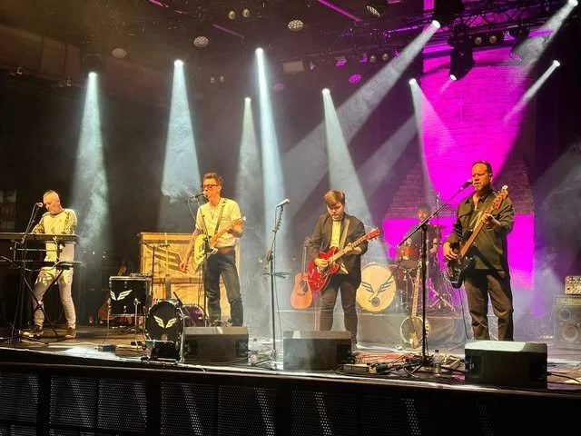 A band of five musicians performing on stage with instruments such as keyboard, electric guitar, and bass guitar, with stage lights and backdrop.