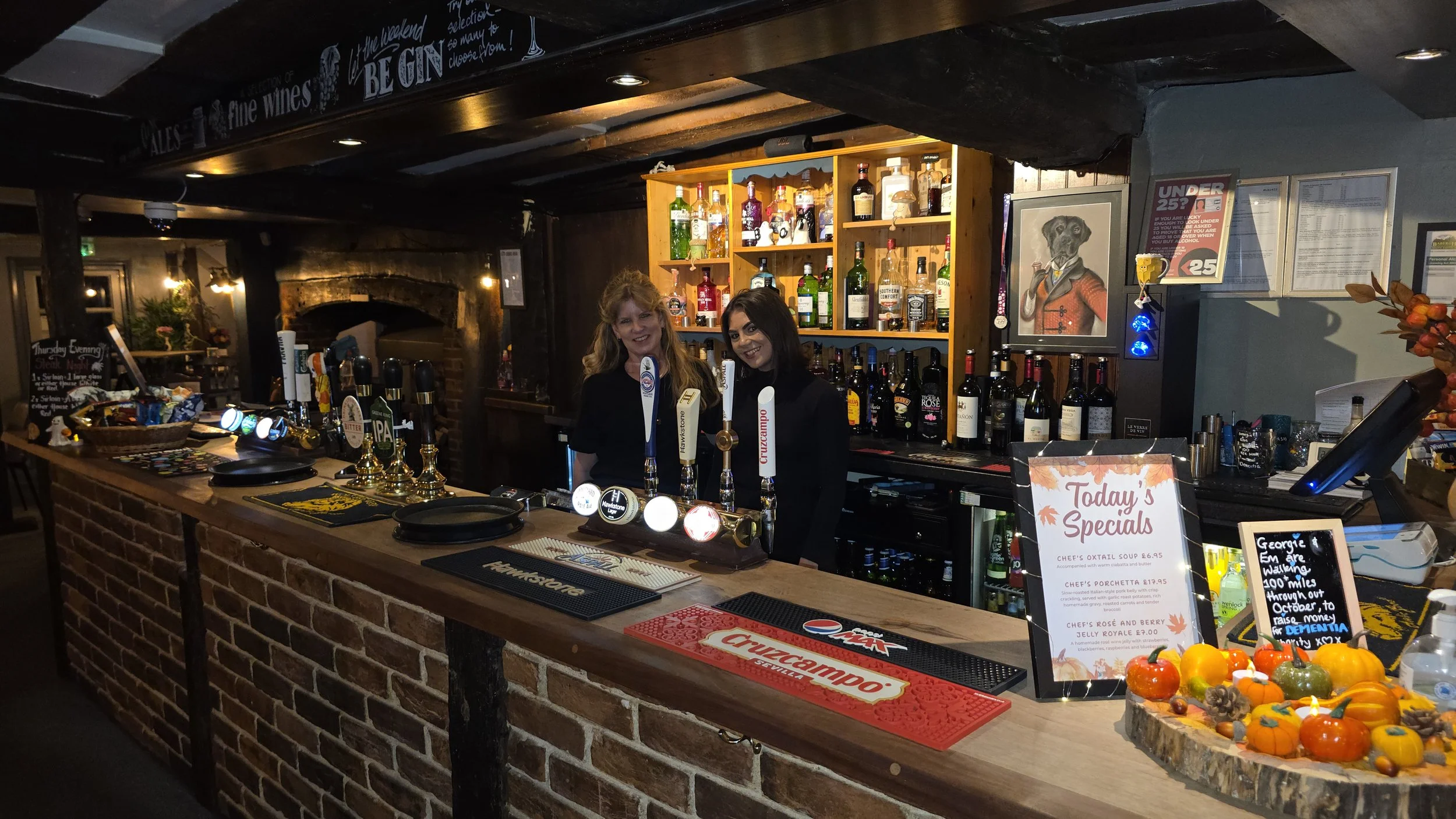 Bar area in a pub with two women behind the counter, shelves of liquor bottles, a menu sign titled 'Today's Specials', and decorated treats on the counter.