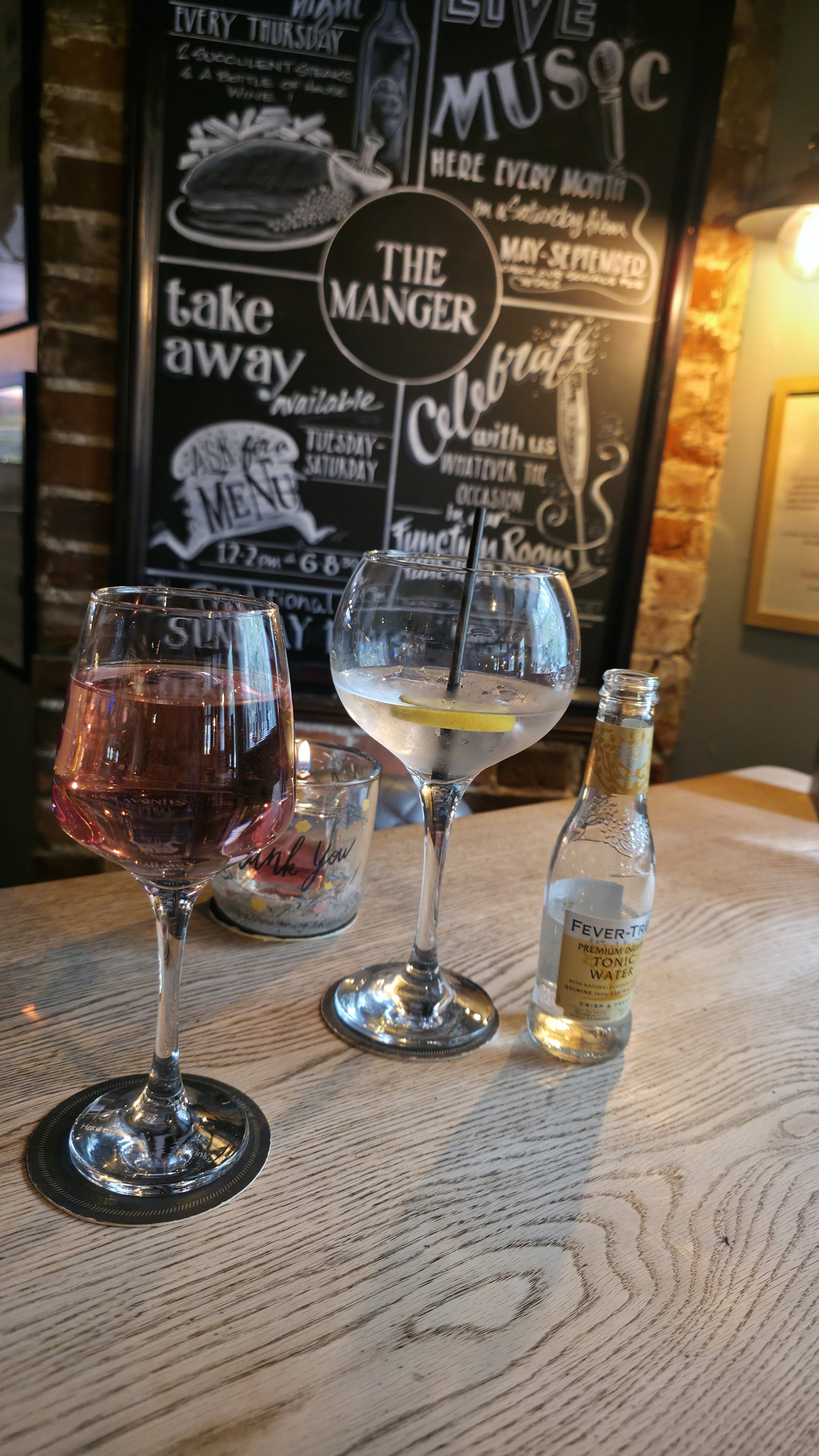 Two glasses of wine and a bottle of tonic water on a wooden table in a bar or restaurant setting. In the background, there is a chalkboard menu with handwritten text, some of which mention music nights and takeout options.
