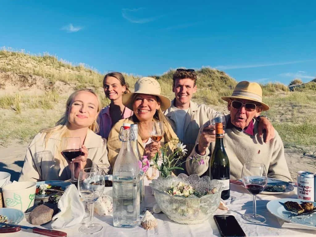 En familie sidder ved et bord udendørs, nyder vin og mad på en solrig dag med sandklitter i baggrunden.