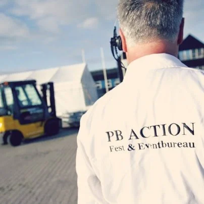 Person med PB ACTION Fest & Eventbureau-logo set fra ryggen, baggrund med telt og gaffeltruck