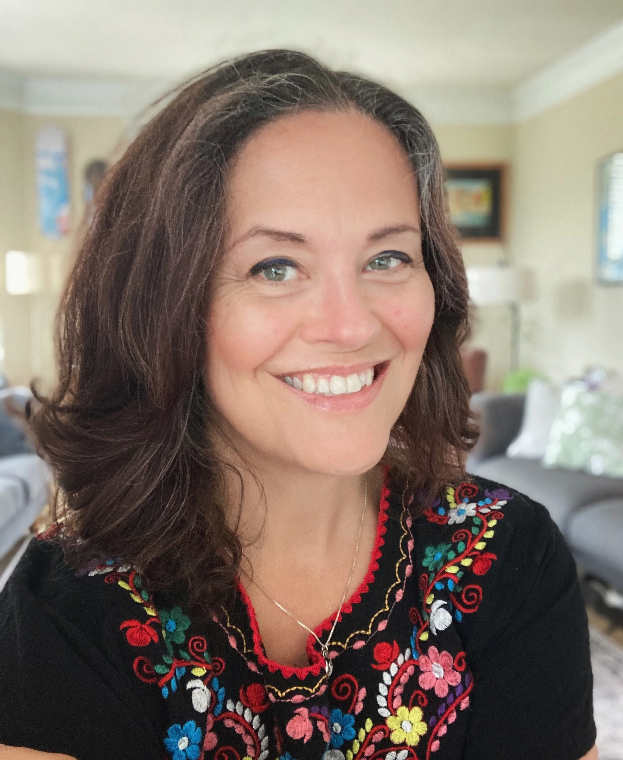 A woman smiling in a room with beige walls. She has shoulder-length brown hair and is wearing a black embroidered top with colorful floral patterns and red trim. She has a silver necklace with a small pendant.