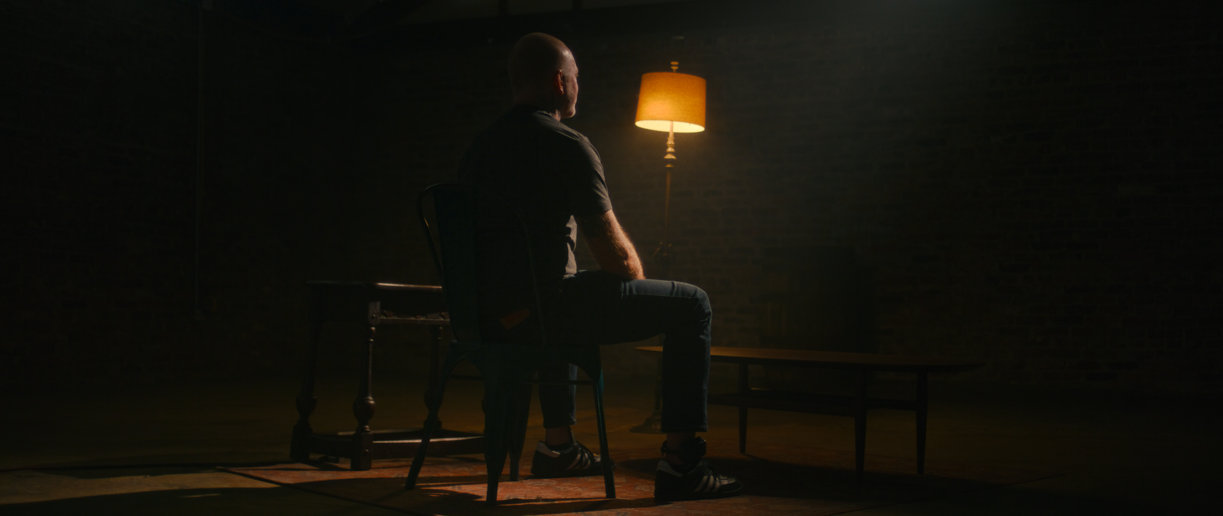 A man with a bald head sitting on a wooden chair in a dimly lit room, facing a table with a small lamp emitting warm light, creating a moody atmosphere.