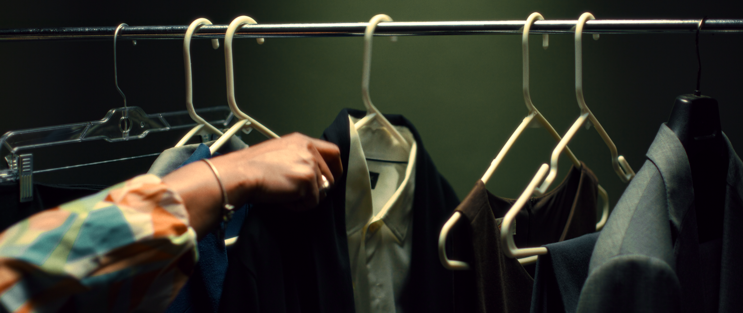 Clothes hanging on a metal rod, including shirts, jackets, and pants, with a person's hand reaching for a black garment.