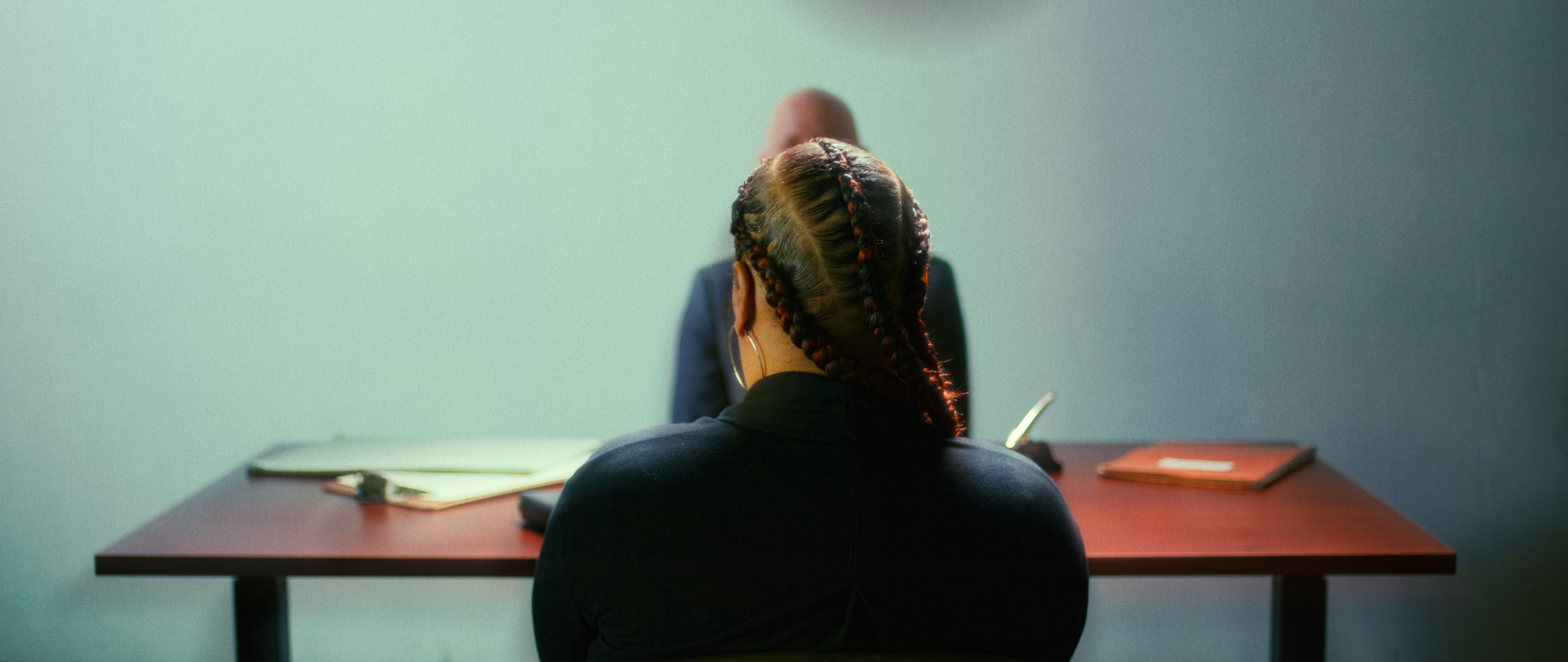 Back of a woman with braided hair sitting at a desk, facing a man in a blue suit, in a sparse office setting.